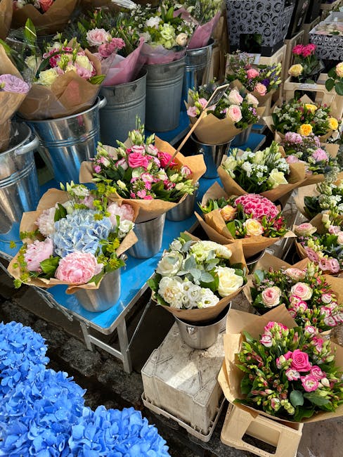 A vibrant display of fresh floral bouquets arranged in metal buckets and wrapped in brown craft paper, featuring a variety of flowers such as pink roses, white and pink peonies, pale blue hydrangeas, 