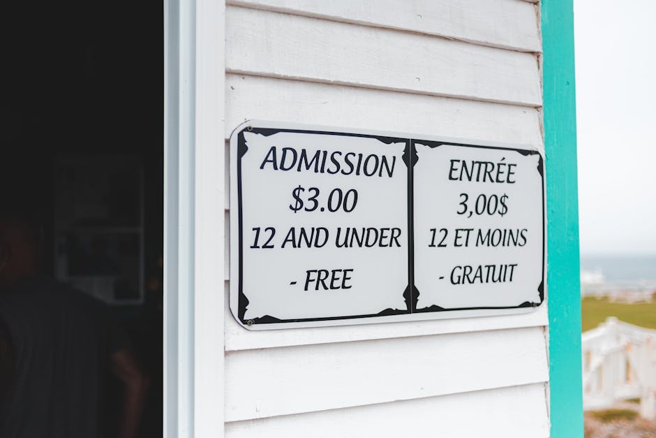 A black and white sign mounted on a white wooden wall displays admission details for an event or venue, with the text 'ADMISSION $3.00 12 AND UNDER - FREE' on the left and 'ENTRÉE 3,00$ 12 ET MOINS -