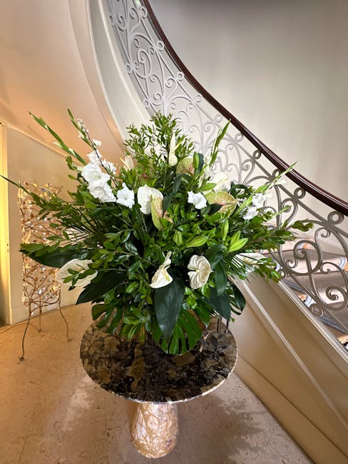 A large, elegant floral arrangement featuring white calla lilies, anthuriums, and various green foliage, including broad leaves and delicate stems. The bouquet is placed in a dark-colored, glossy vase