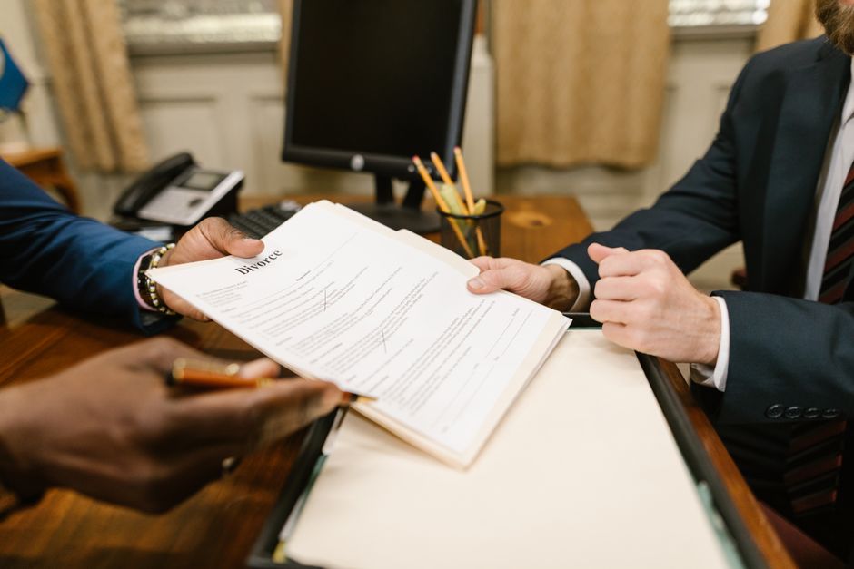 A close-up of a person in a dark suit and white shirt holding a legal divorce document, with visible text and signatures, as another individual in a blue blazer holds a pen. The scene is set at a wood