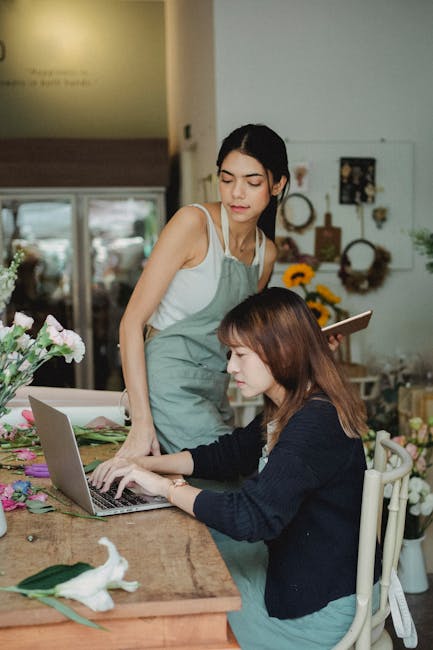 A florist from the company is instructing a customer at a flower shop, with various fresh flowers in shades of pink, purple, and white arranged on the wooden table. The florist, wearing a light green