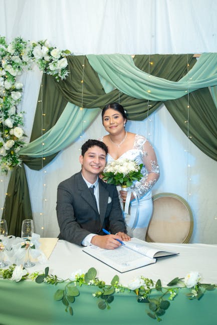 A young man in a dark suit and white shirt is seated at a table, smiling as he signs a document, while a young woman in a white lace wedding dress stands behind him, holding a bridal bouquet of white
