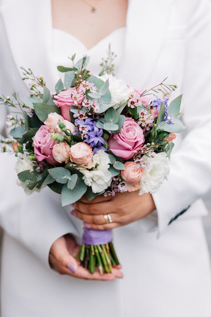 A person dressed in a white blazer holding a fresh floral bouquet with both hands. The bouquet features pink roses, pale peach ranunculus, white lisianthus, and sprigs of purple and white accents, all