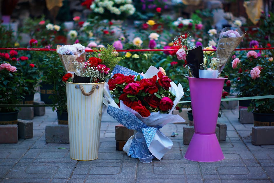A floral arrangement featuring a bouquet of red roses wrapped in white and blue patterned paper, placed between two vases on a paved outdoor area. To the left, a white cylindrical vase with beige vert
