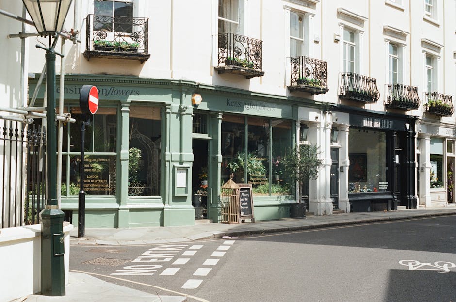 A row of quaint flower shops on a street corner with large display windows filled with fresh floral arrangements, including roses, lilies, and daisies in various bright colors. The shop fronts are pai