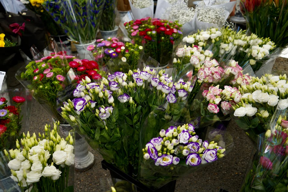 A variety of fresh floral arrangements displayed on a table, featuring delicate purple and white eustoma flowers, pink and white roses, and clusters of small, vibrant pink and purple daisies. The flow
